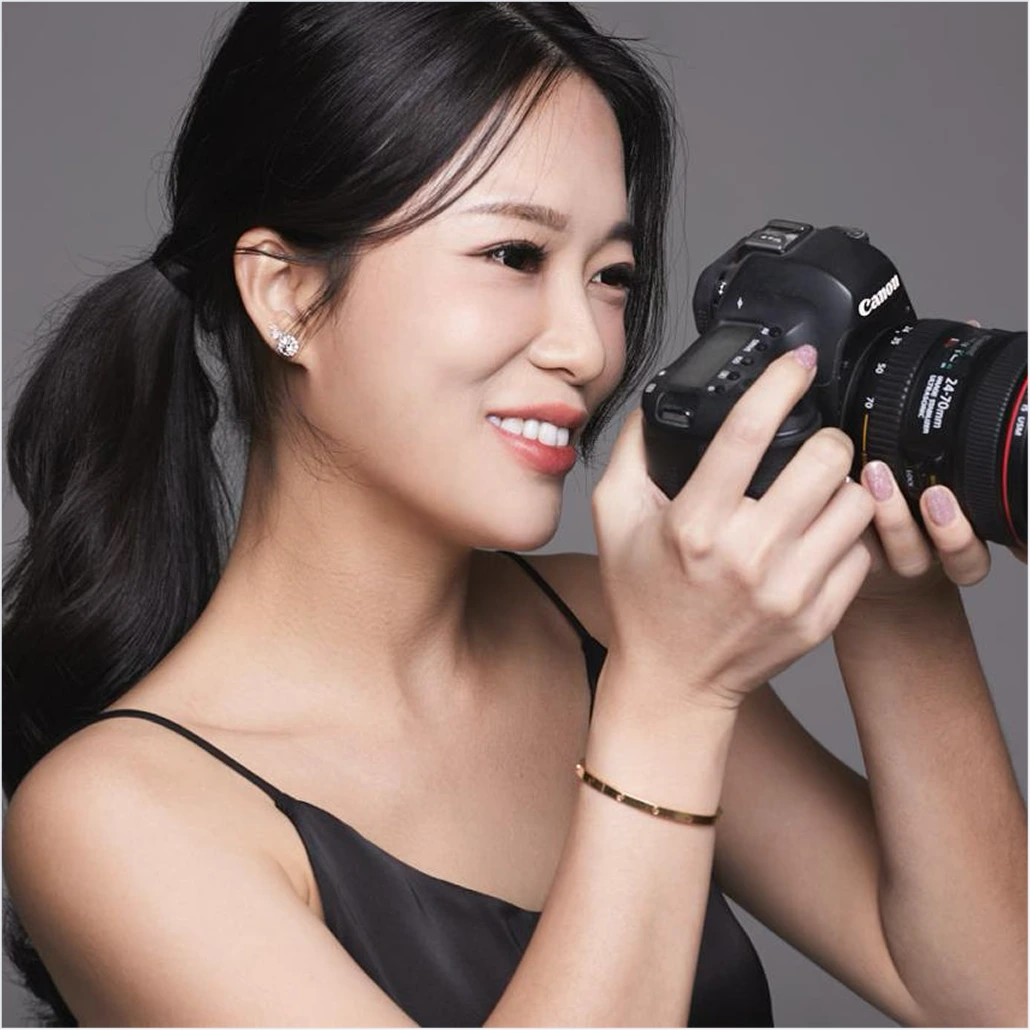 Stephanie Teo raising a Canon DSLR to her eye, smiling behind the viewfinder